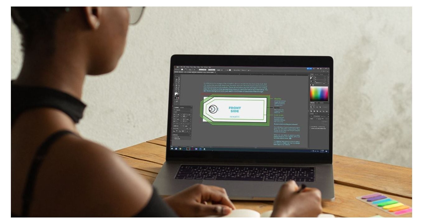 A woman sitting at the desk looking at the laptop screen with Adobe Illustrator print template opened.