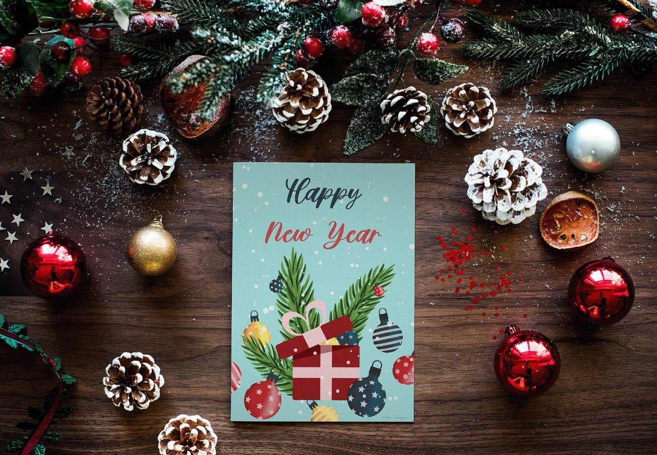 Christmas card on a wooden desk surrounded with ornaments, pinecones and branches.