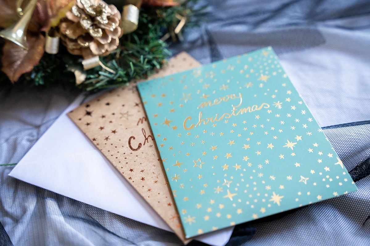 Two holidays cards with gold foil details and and envelope next to Christmas decorations.