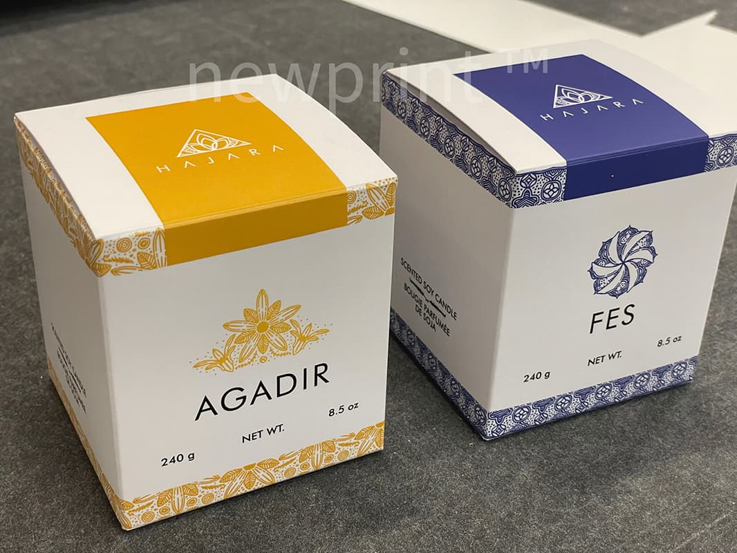 Two different paperboard candle boxes next to each other, both white, one with yellow and the other with blue design elements.